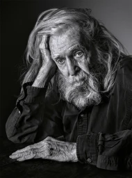 Close-up monochrome portrait of an elderly man with long hair and a full beard, resting his head on his hand.