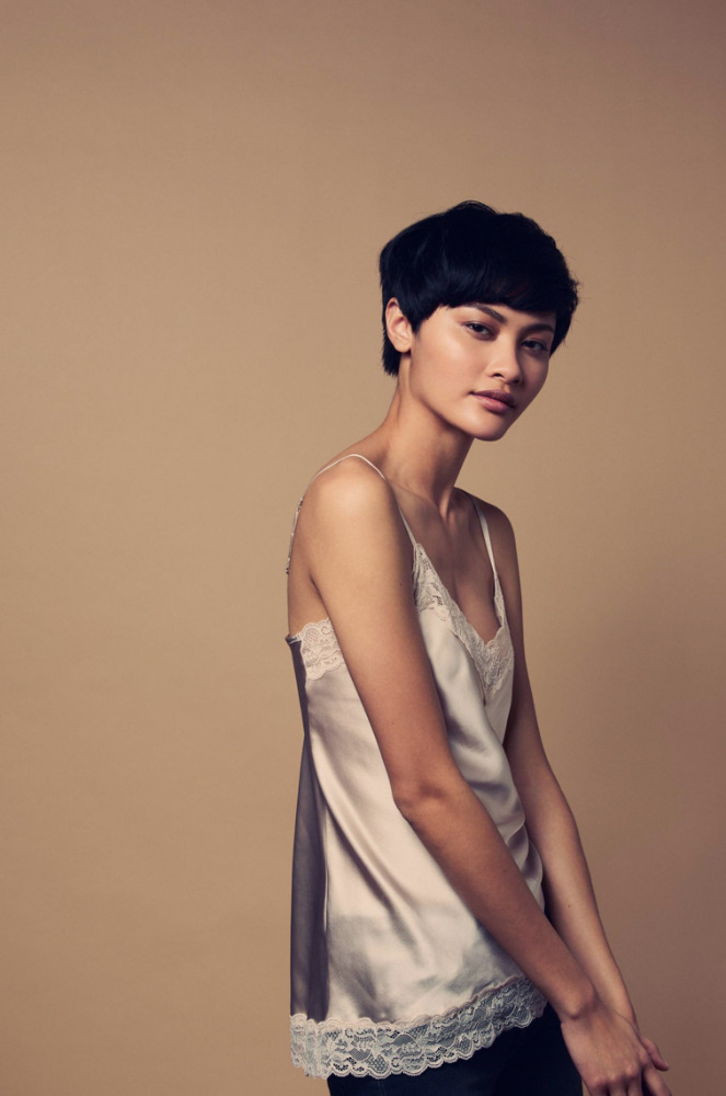 Portrait of a young woman with short dark hair wearing a lace-trimmed ivory satin camisole against a neutral background in soft studio lighting.