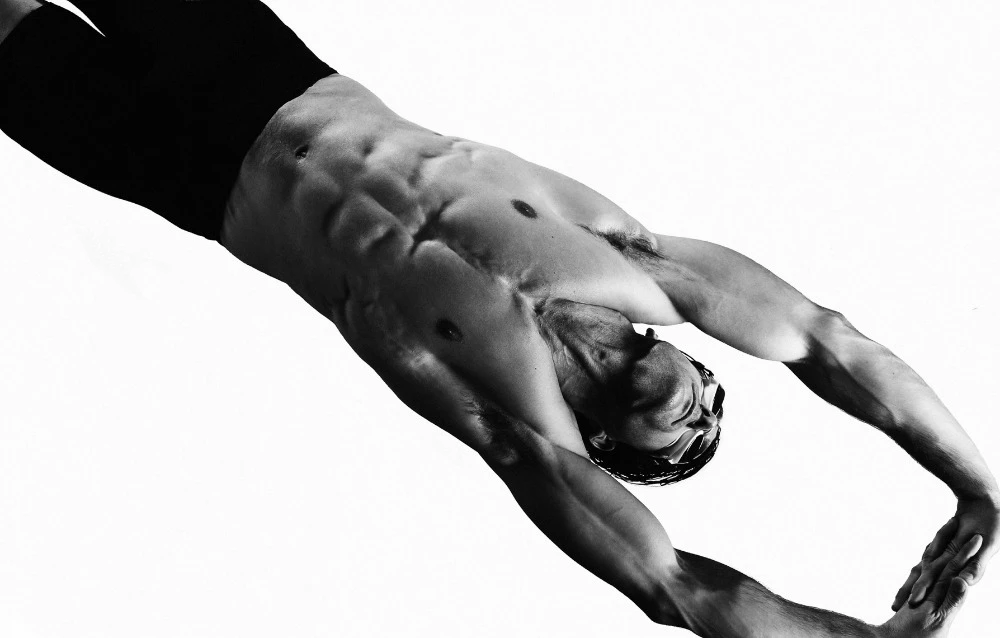 Athletic male swimmer in black swimsuit dives with outstretched arms in black and white, highlighting muscular physique against a white background.