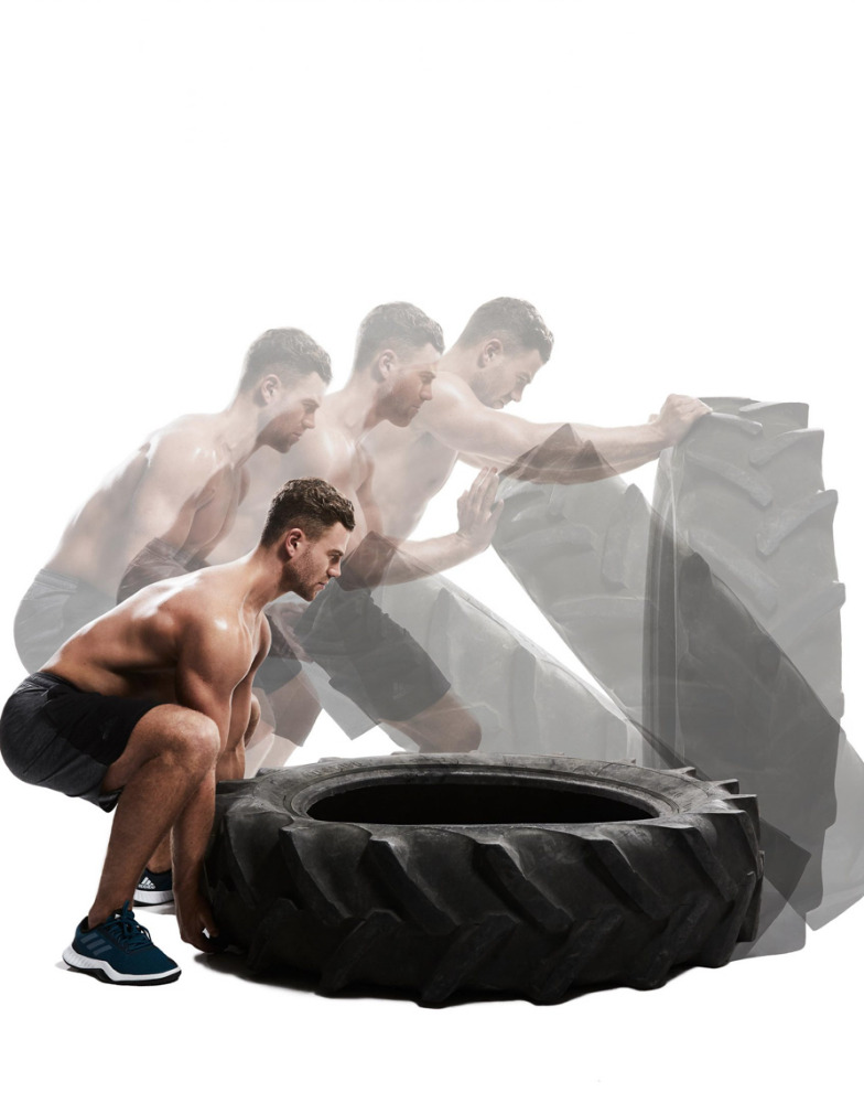 Shirtless man performs tire flip workout in studio setting, exhibiting muscular build and athletic strength during exercise routine.