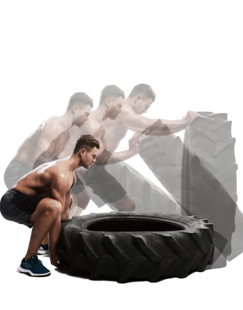 Shirtless man performs tire flip workout in studio setting, exhibiting muscular build and athletic strength during exercise routine.