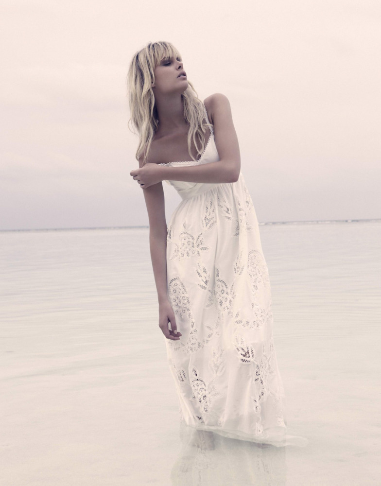 Fashion shot of a blonde woman in a long white dress standing in shallow water on a cloudy day, looking up and away from the camera.