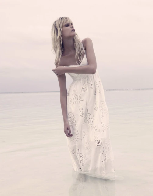 Fashion shot of a blonde woman in a long white dress standing in shallow water on a cloudy day, looking up and away from the camera.