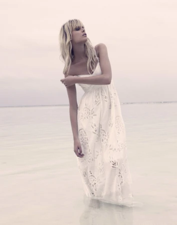 Fashion shot of a blonde woman in a long white dress standing in shallow water on a cloudy day, looking up and away from the camera.