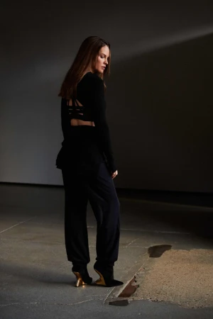 A woman in a black strappy top and trousers with sculpted gold-heeled shoes stands on a concrete floor in a moody interior space.