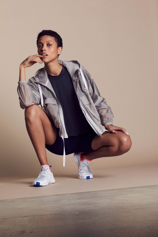 Woman in athletic wear poses crouching indoors in front of a neutral background, showcasing a lightweight jacket, shirt, shorts, and running shoes.