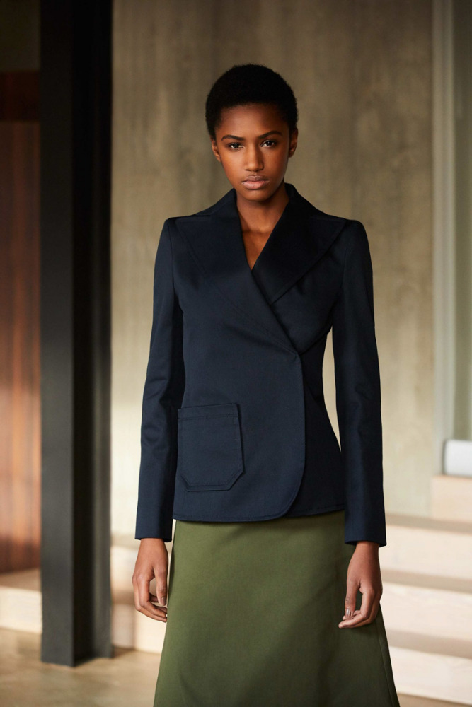 Indoor portrait of a Black female model in a navy blazer and green skirt against a neutral studio backdrop in a fashion editorial.