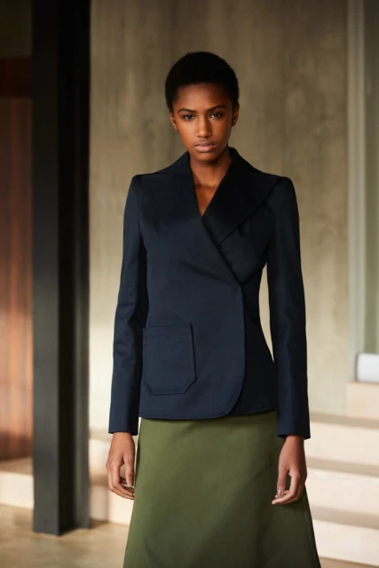 Indoor portrait of a Black female model in a navy blazer and green skirt against a neutral studio backdrop in a fashion editorial.