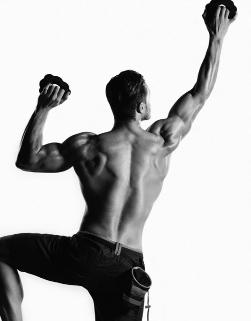 Black and white shot of a muscular adult male climbing a rock wall indoors, arms extended, wearing dark athletic shorts and climbing gear.