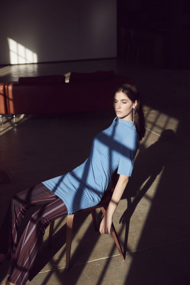 Adult woman in blue shirt and striped pants sits on a chair in a room with light streaming through the windows, casting shadows.