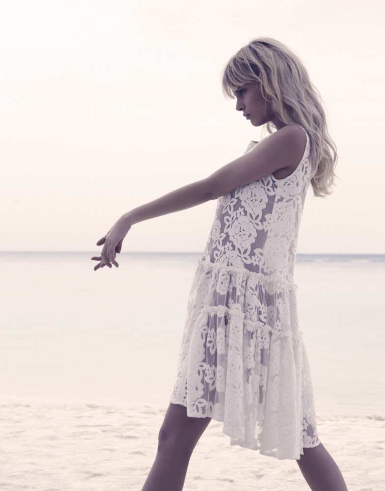 A blonde model wearing a white lace dress posing gracefully on a sandy beach under an overcast sky.