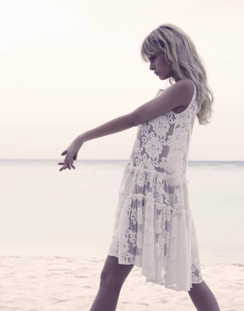 A blonde model wearing a white lace dress posing gracefully on a sandy beach under an overcast sky.