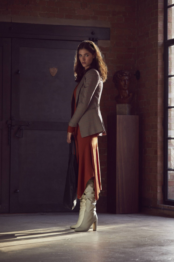 Full-length shot of a woman in a rust-colored dress, beige blazer, and white heeled boots, posing in an indoor location with neutral tones.