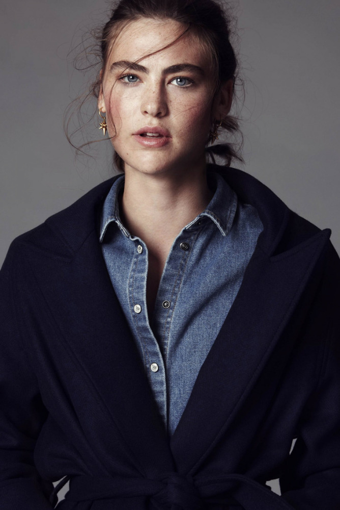 Close-up of a fair-skinned adult female model with blue eyes and freckles, wearing a denim shirt and blue trench coat against gray.