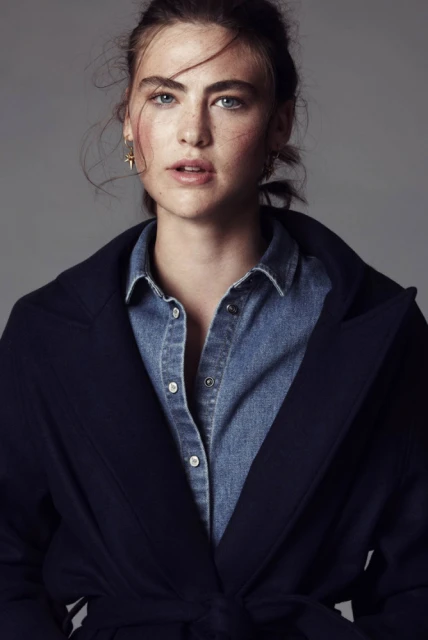 Close-up of a fair-skinned adult female model with blue eyes and freckles, wearing a denim shirt and blue trench coat against gray.