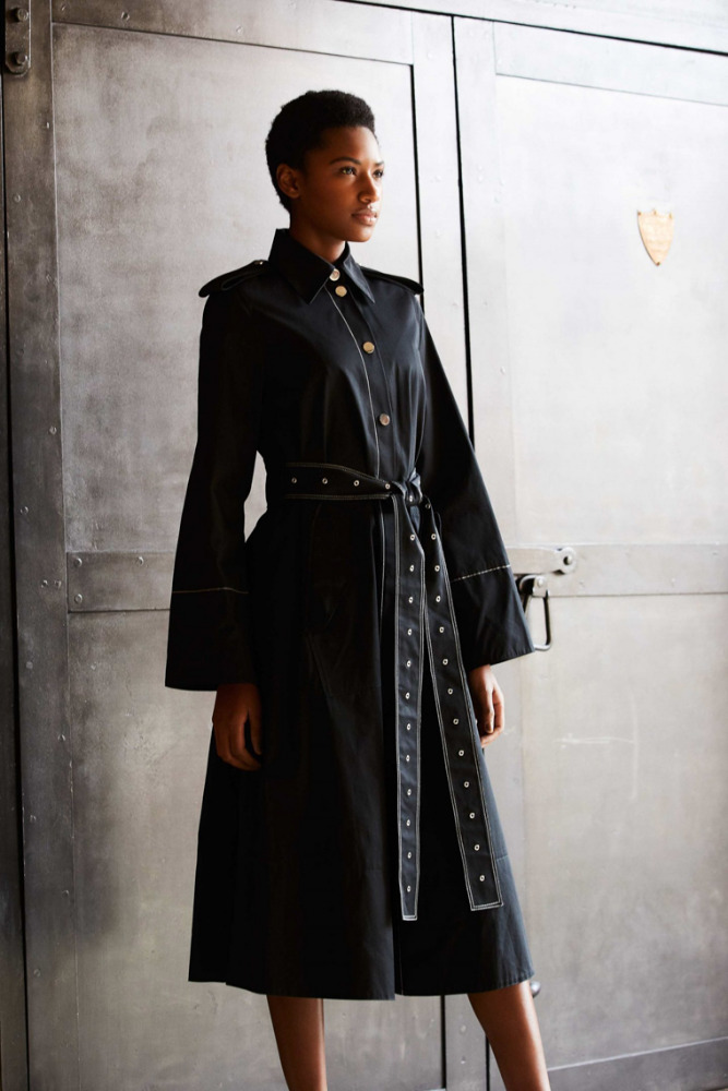 Woman in a fashionable black trench coat with bell sleeves and studded belt poses against a textured metal background in a studio shot.