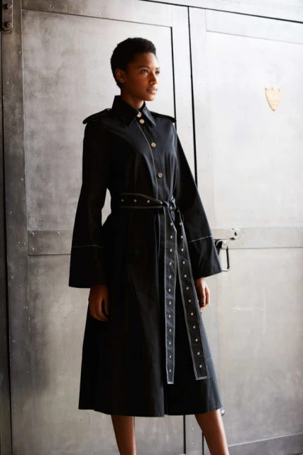 Woman in a fashionable black trench coat with bell sleeves and studded belt poses against a textured metal background in a studio shot.