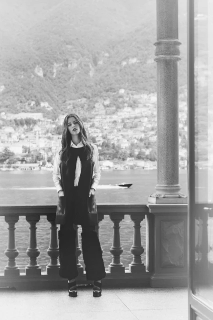 Full shot of a female fashion model in a dark jumpsuit with a white blouse standing on a balcony overlooking a lake and mountains.