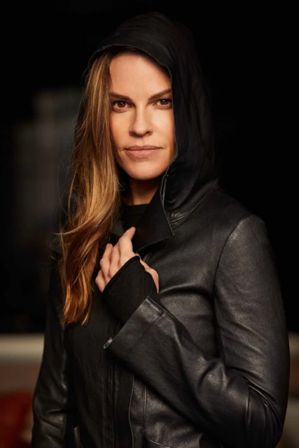 Portrait of adult female actress Hilary Swank with long hair, wearing a black hooded leather jacket against a dark background.
