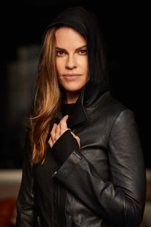 Portrait of adult female actress Hilary Swank with long hair, wearing a black hooded leather jacket against a dark background.