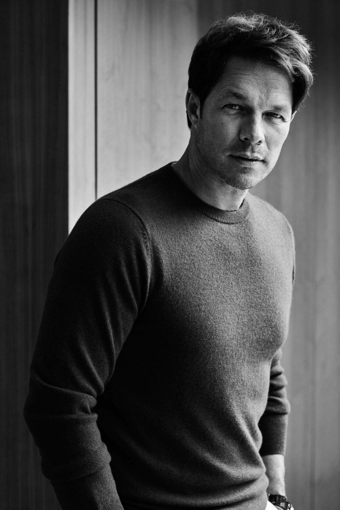 Monochrome portrait of a fair-skinned adult male in a sweater, posing indoors with a neutral expression. The lighting is soft and diffused.