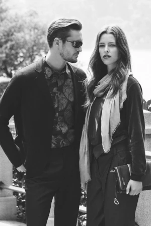 Black and white portrait of a man in sunglasses and patterned shirt standing beside a woman wearing a scarf and holding a clutch
