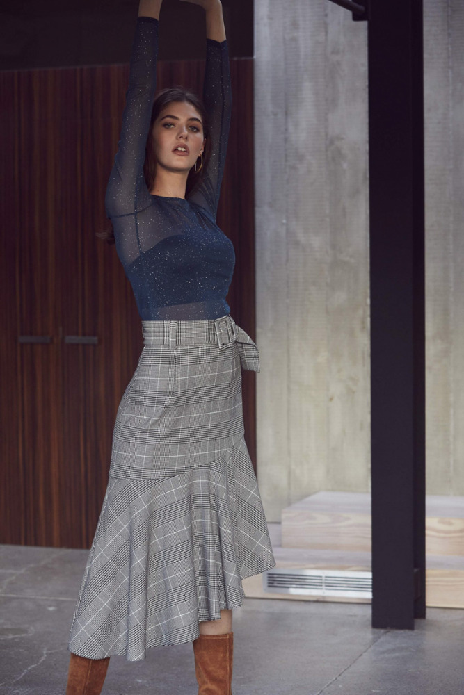 A woman poses in a blue top and plaid skirt in a modern indoor setting, showcasing a high-fashion editorial style and elegant design.