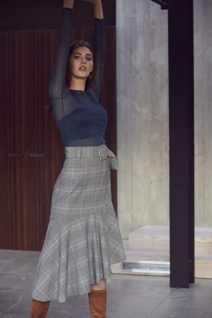 A woman poses in a blue top and plaid skirt in a modern indoor setting, showcasing a high-fashion editorial style and elegant design.