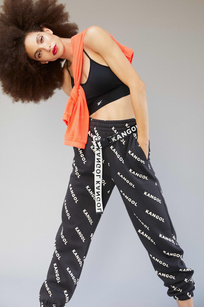 A woman in a black sports bra and Kangol pants poses against a grey background, an orange sweater draped over her shoulders.
