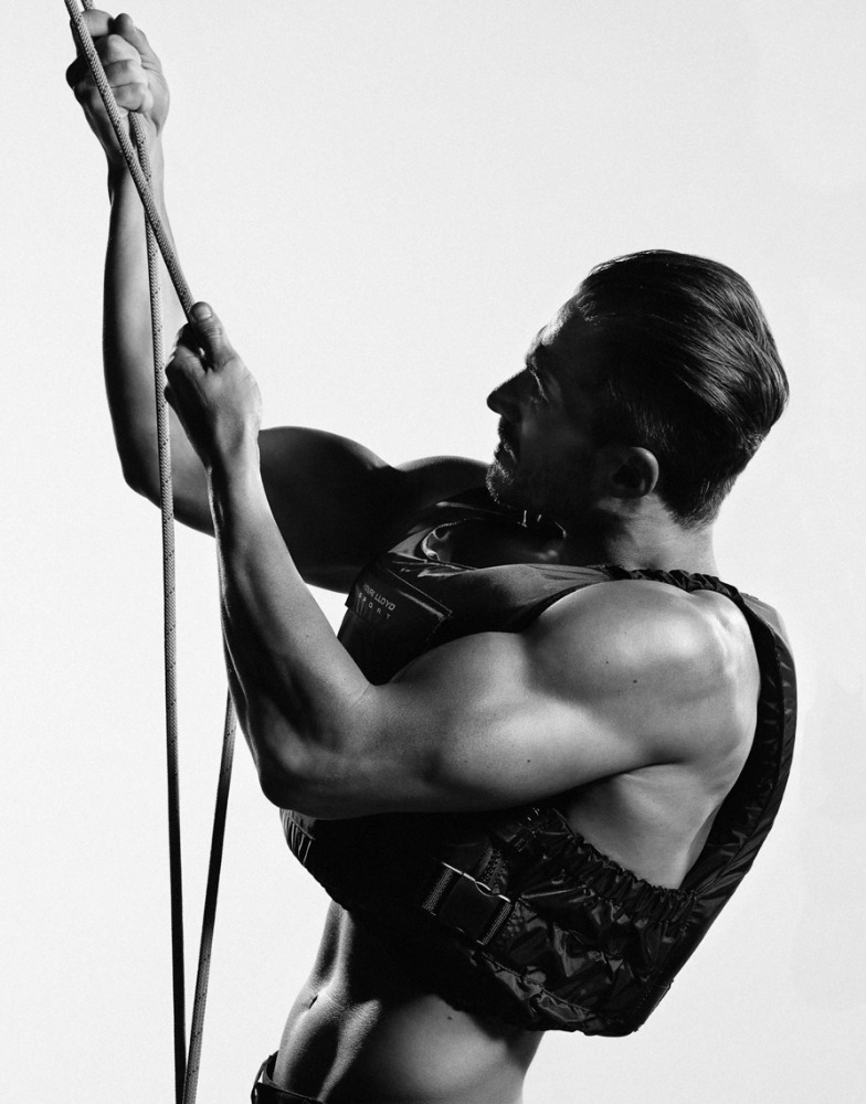 Monochrome photo of an adult male pulling on a rope, wearing a vest; close-up on torso and face in an indoor environment against a white backdrop.