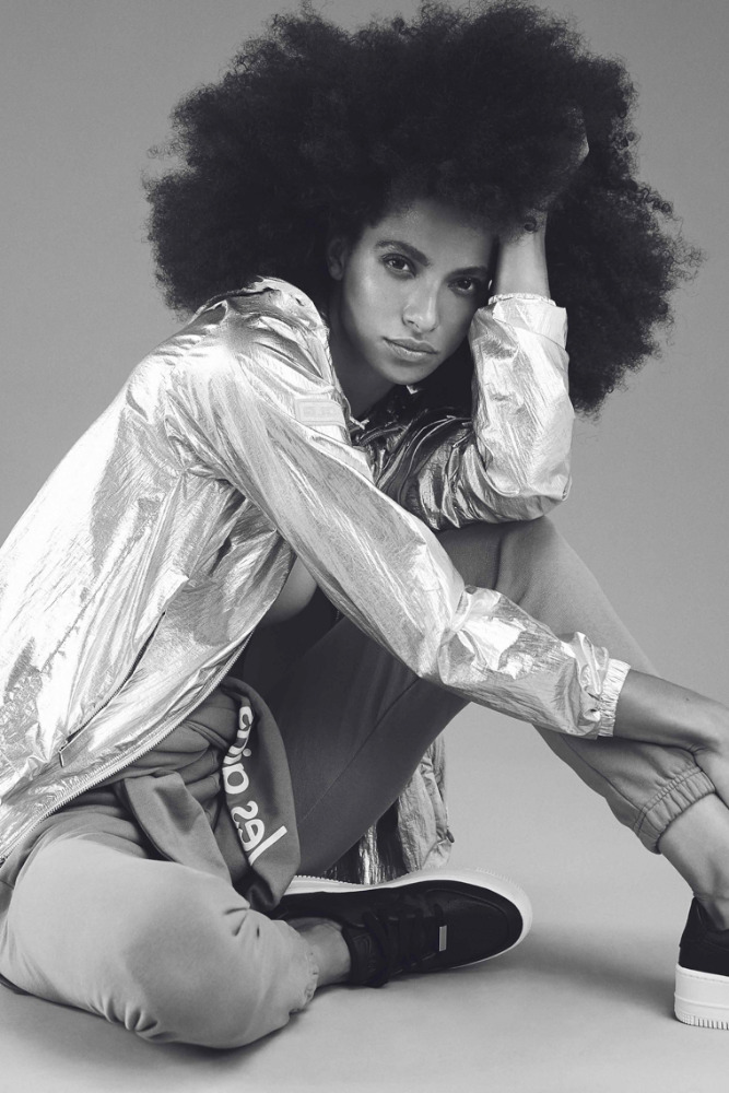 Black and white portrait of a woman with an afro hairstyle wearing a metallic jacket, seated indoors with neutral backdrop.