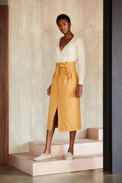 A slender Black woman wearing a cream wrap sweater and gold midi skirt stands on steps in an indoor fashion shot.
