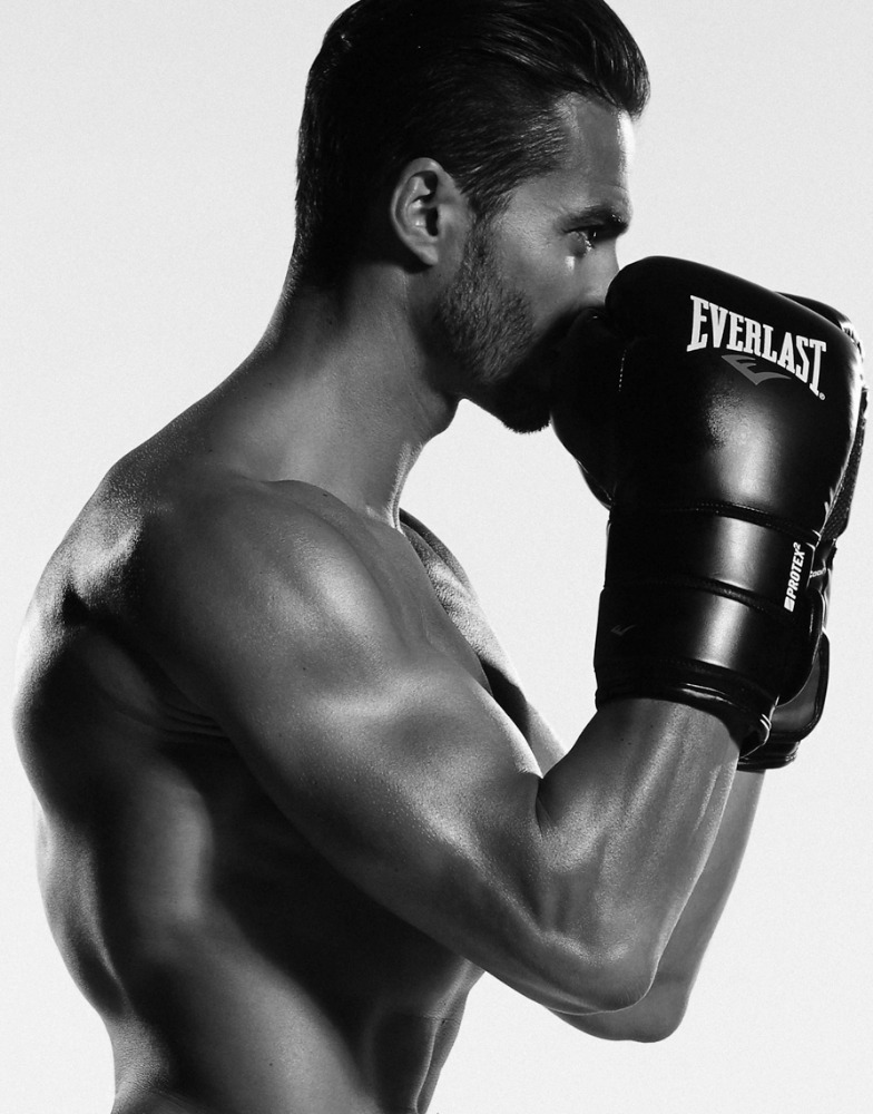 Black and white studio shot of a shirtless man in boxing gloves with light stubble posing against a white background.
