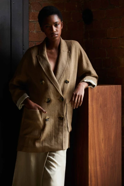 A dark-skinned woman poses in a tan double-breasted jacket and cream skirt against a brick wall, bathed in studio light.
