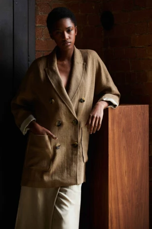 A dark-skinned woman poses in a tan double-breasted jacket and cream skirt against a brick wall, bathed in studio light.