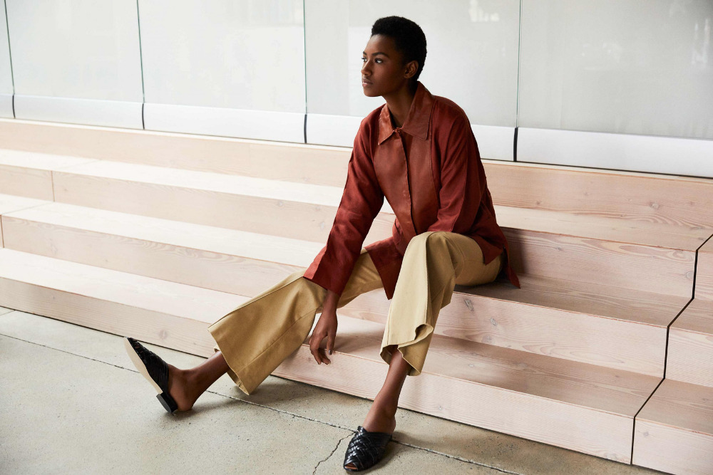 An adult woman sits on wooden stairs in an auburn shirt jacket and light khaki pants, complemented by black shoes, against a white wall.