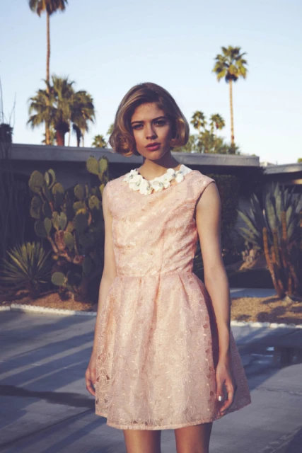 A young woman poses in a peach-colored lace dress with a white flower collar in front of a modern house and palm trees outdoors.