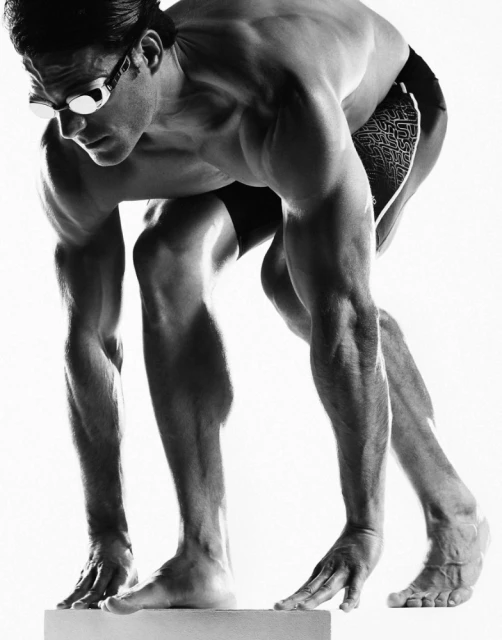 Black and white photo of a swimmer in a crouched position wearing goggles, preparing to dive from a platform in a bright studio.
