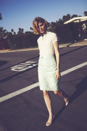 Woman with short blonde hair wearing a white short-sleeve top and pale green lace skirt standing on an asphalt street.