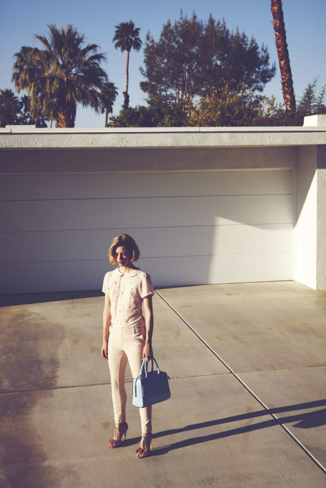 A woman in a light-colored jumpsuit stands in front of a white building with palm trees visible in the background on a sunny day.