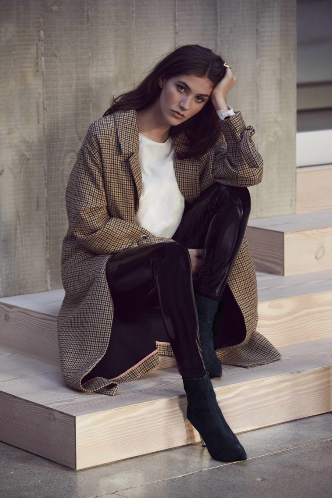 Woman wearing a patterned tweed coat and shiny black trousers sitting on wooden steps outdoors.