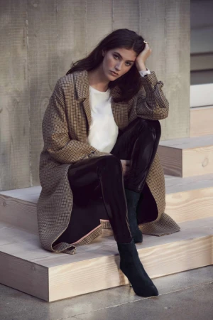 Woman wearing a patterned tweed coat and shiny black trousers sitting on wooden steps outdoors.