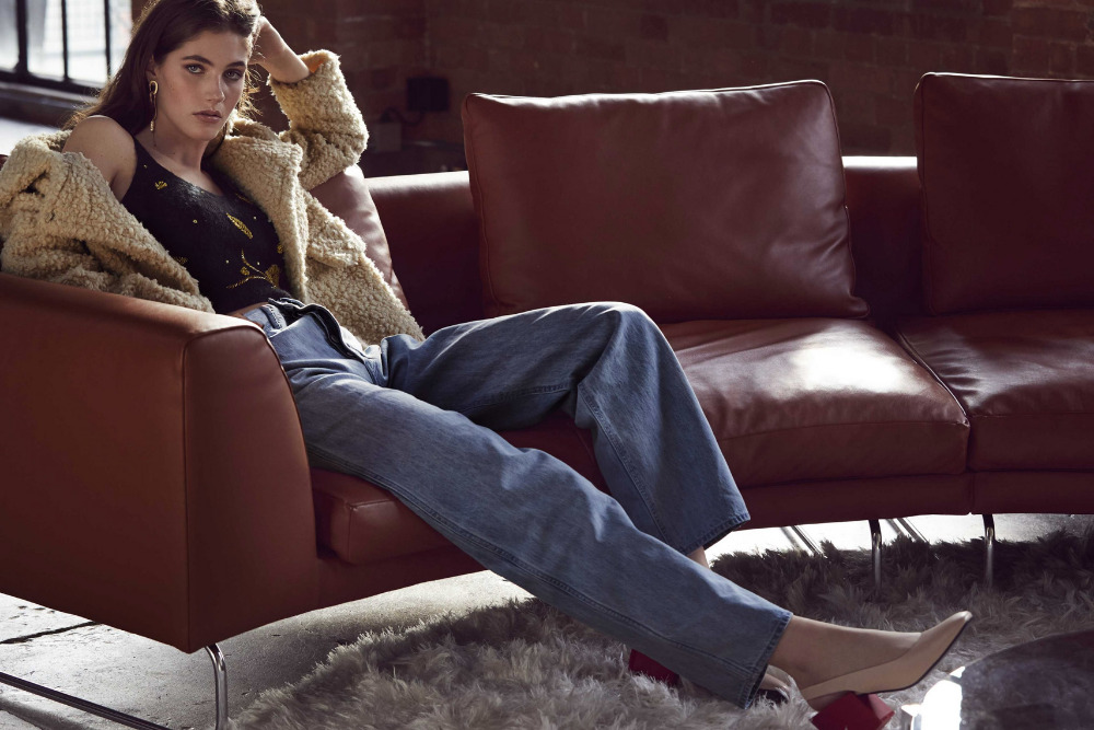Stylish woman relaxes on a rust-colored leather couch wearing jeans, a black tank top, and a sherpa jacket in a casual indoor setting.
