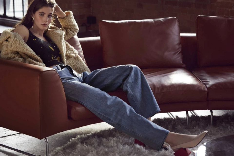 Stylish woman relaxes on a rust-colored leather couch wearing jeans, a black tank top, and a sherpa jacket in a casual indoor setting.