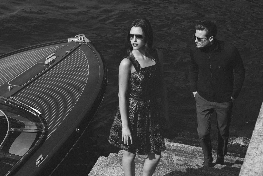 Black and white image of a stylish couple posing by a sleek speedboat on a waterfront. The woman wears a dress, the man a jacket and trousers.
