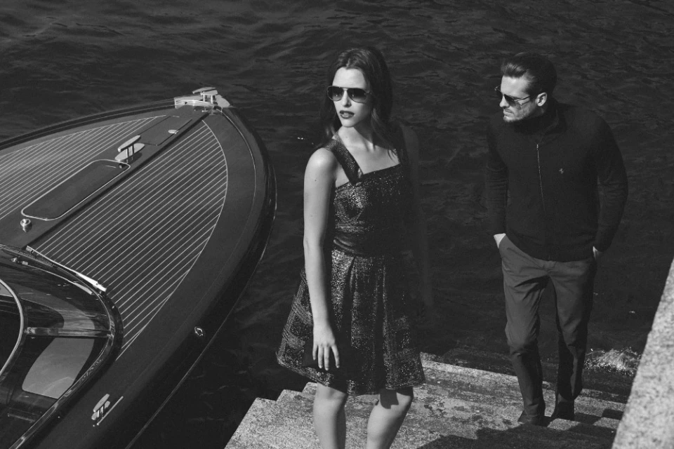 Black and white image of a stylish couple posing by a sleek speedboat on a waterfront. The woman wears a dress, the man a jacket and trousers.