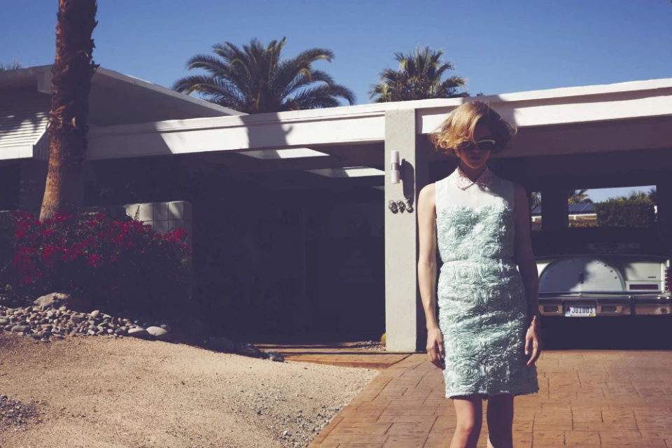 A woman in a patterned dress stands outside a modern house in bright sunlight, with palm trees and an open carport.