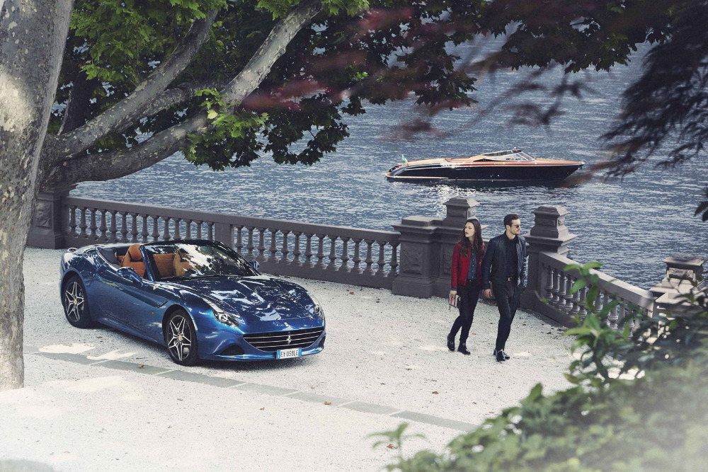 Blue Ferrari convertible car parked near a stone fence, with a couple walking along the water and a speedboat visible on the lake.