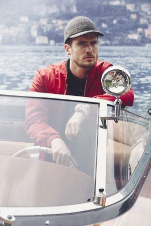 Man wearing a grey tweed cap and red jacket leaning on the windshield of a boat on a lake by Ben Riggot