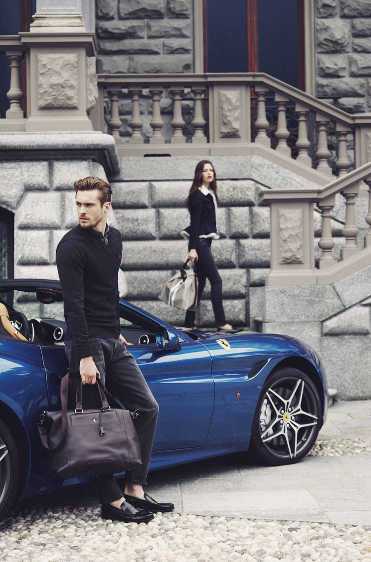 Well-dressed man with leather bag poses near a blue convertible car with a woman on a stone staircase in the background.